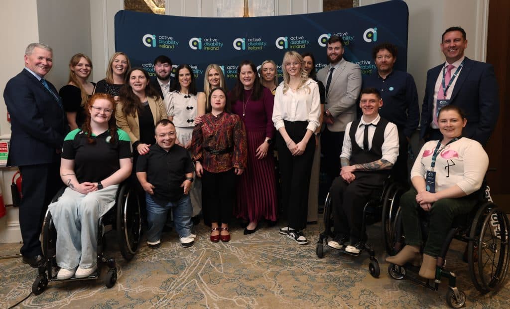 A large group of adults pose together for a group photo in front of an “Active Disability Ireland” backdrop. The group includes people standing and people using wheelchairs. They are smiling and dressed in a mix of formal and casual clothing, at a conference setting.