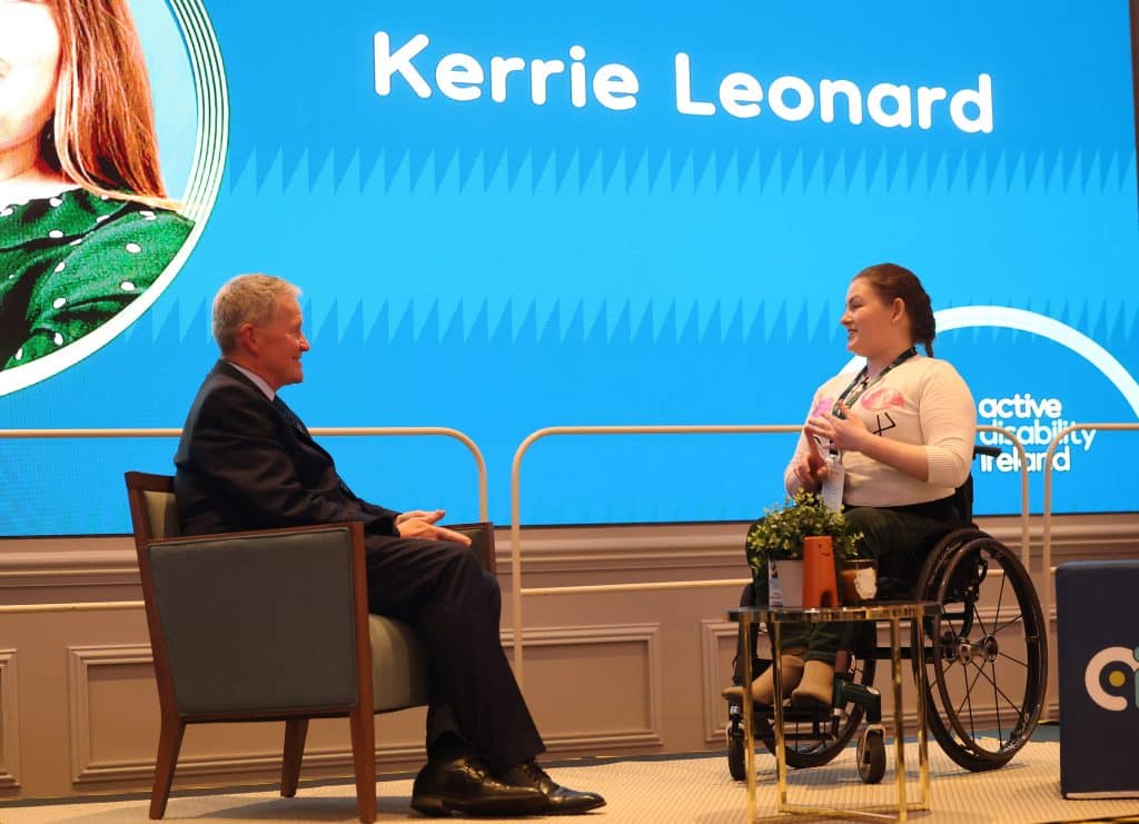 Image of wheelchair user Kerrie Leonard speaking to Rob Hartnett who is seated on an armchair on a stage.